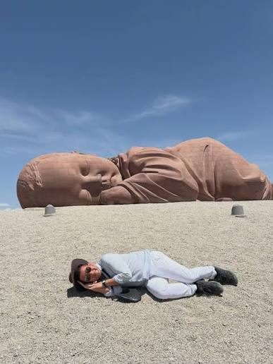 Person resting beneath a large reclining Buddha sculpture in an open landscape