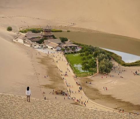 A desert oasis with water, trees and a pavilion viewed from a sand dune